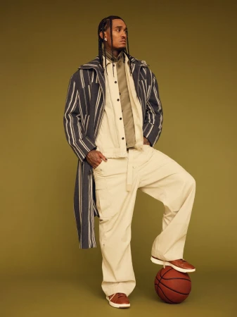 Man in fashionable striped overcoat, tan pants, brown shoes posing with foot on basketball in a studio with tan backdrop.