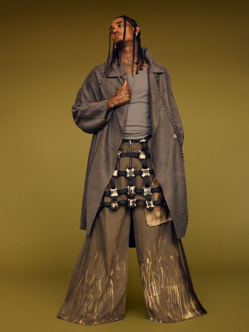 A man in a gray overcoat, tank top, and wide-legged pants stands in a studio with a neutral background, shot at eye level.