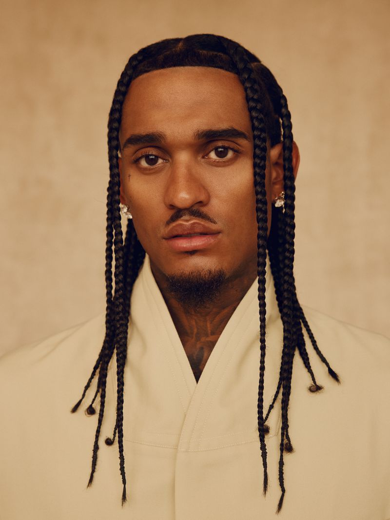Close-up portrait of an adult male with braided hair, wearing an off-white shirt, against a neutral backdrop in soft lighting.