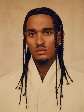 Close-up portrait of an adult male with braided hair, wearing an off-white shirt, against a neutral backdrop in soft lighting.