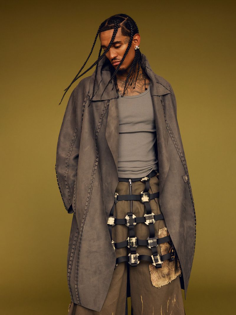 Adult male posing in a photo studio, wearing a grey top, long grey suede coat with a decorative belt, and brown pants.