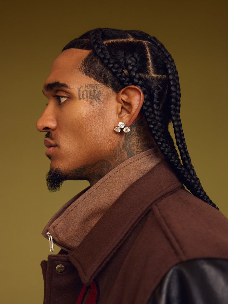 Profile shot of a man with braided hair and a tattoo, wearing a brown jacket, set against a neutral olive background.