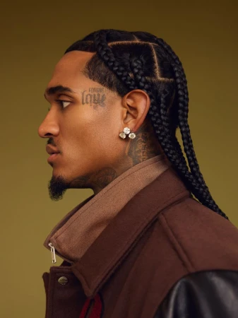 Profile shot of a man with braided hair and a tattoo, wearing a brown jacket, set against a neutral olive background.