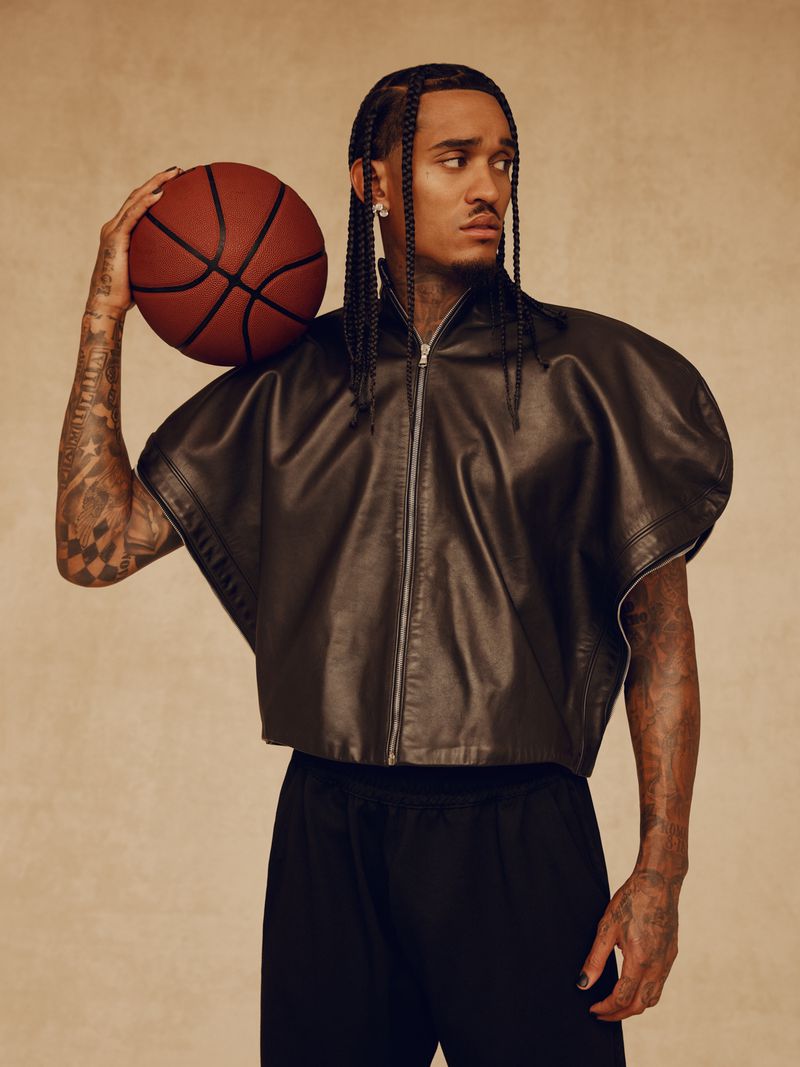 An adult male with braided hair holds a basketball, dressed in a black leather jacket and sweatpants, against a neutral background.