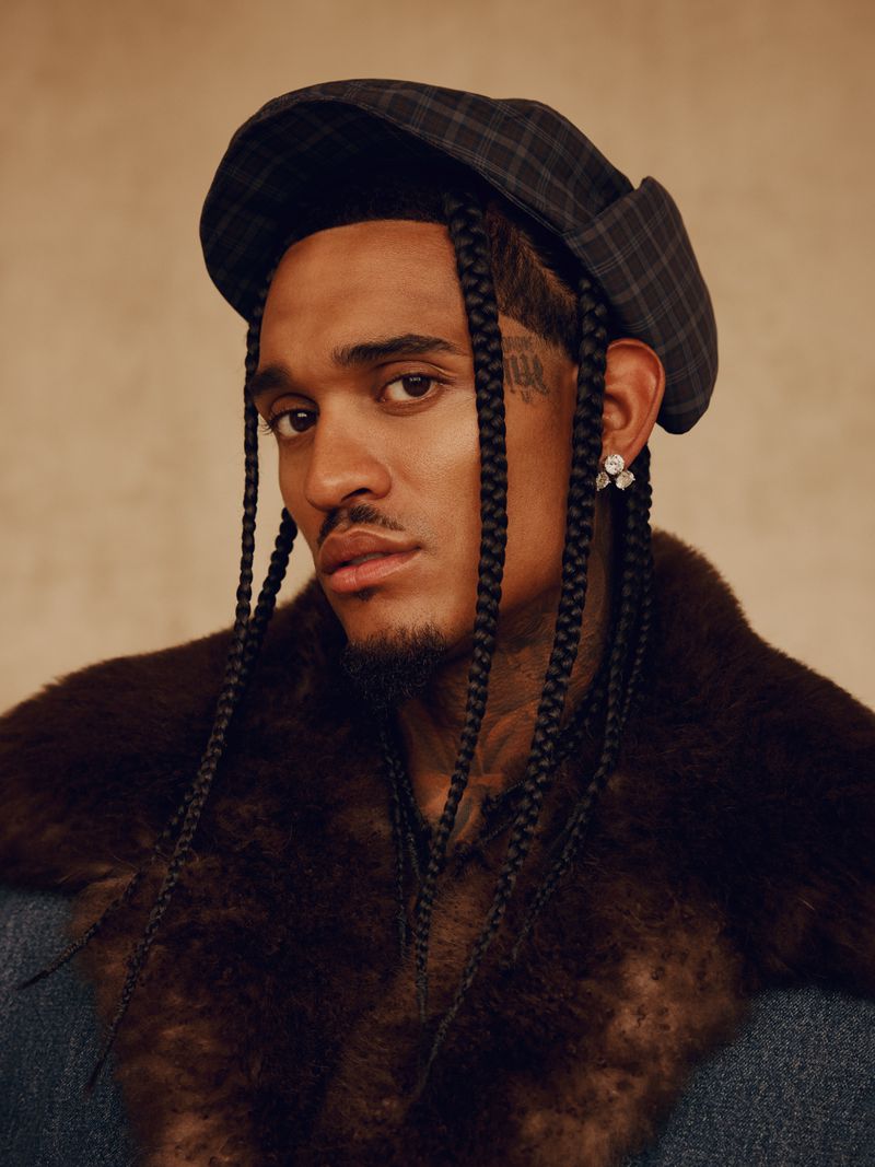 Close-up portrait of a young adult male wearing a plaid hat and a fur collar jacket against a neutral tan-colored background.
