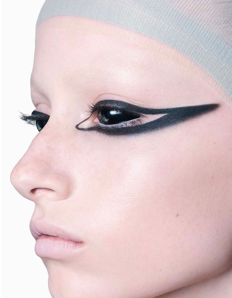 Close-up portrait of a woman with pale skin and striking black eyeliner and black contact lenses against a stark white background.