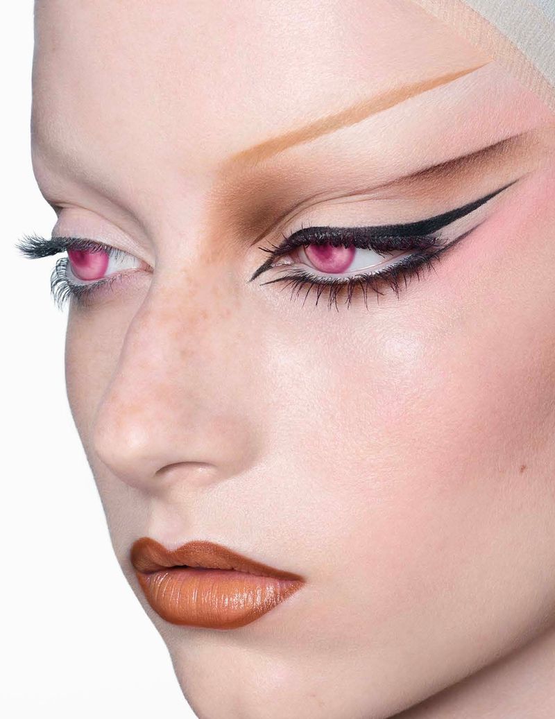 Close-up of a woman's face with dramatic eye makeup, including bold eyeliner and extended eyebrows, and lips with caramel colored lipstick.