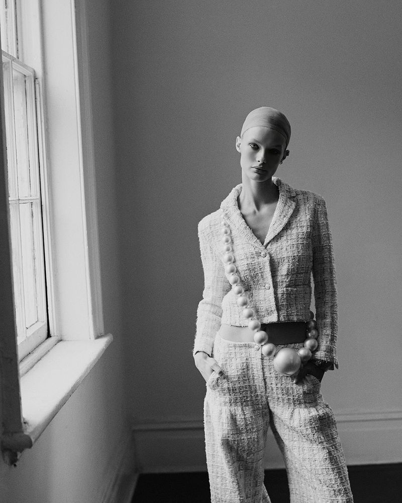 Elegant black-and-white fashion portrait of a slim adult woman in a tweed suit and oversized pearl jewelry, photographed indoors.