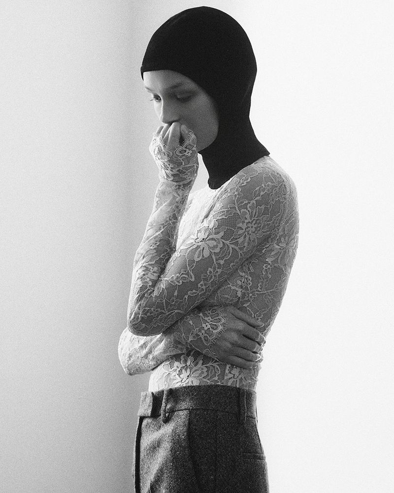 Monochrome portrait of an adult woman wearing a black balaclava and lace top against a plain backdrop in a studio setting.