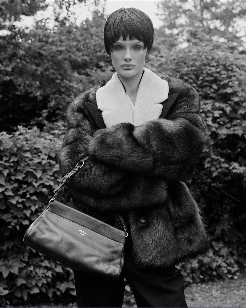 Black-and-white fashion shot of a woman in a fur coat with a Prada handbag, posing outdoors against a backdrop of trees and foliage.