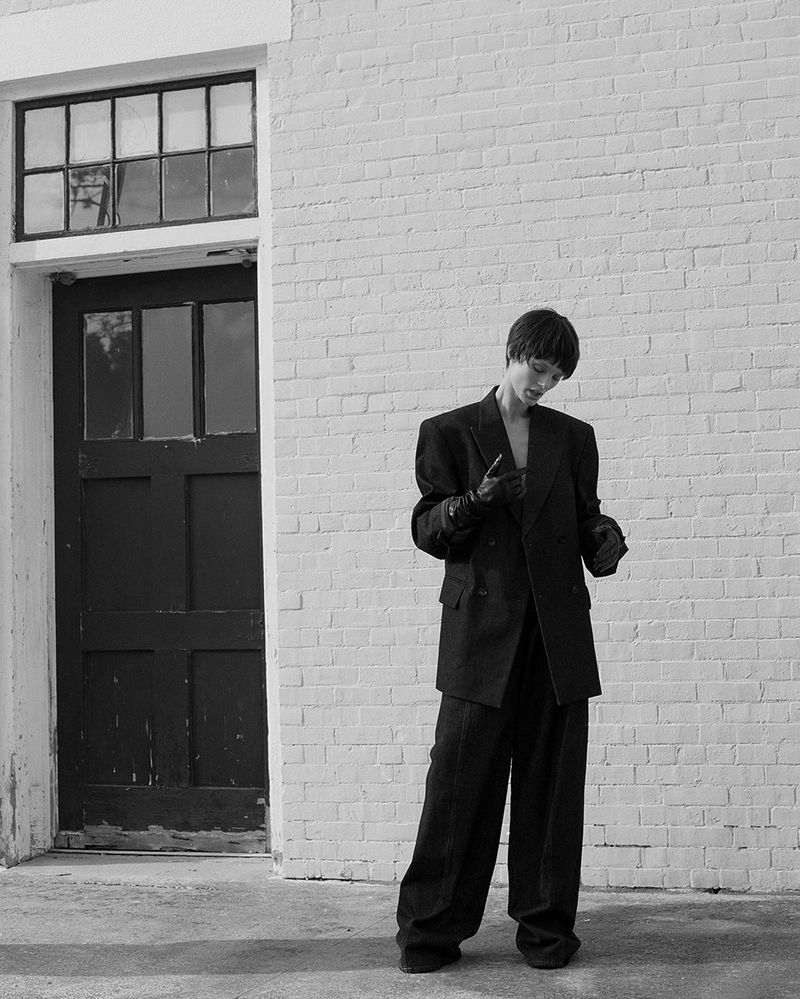 Monochrome image of a woman in a dark suit standing outdoors against a brick wall, looking at a device in her gloved hands.