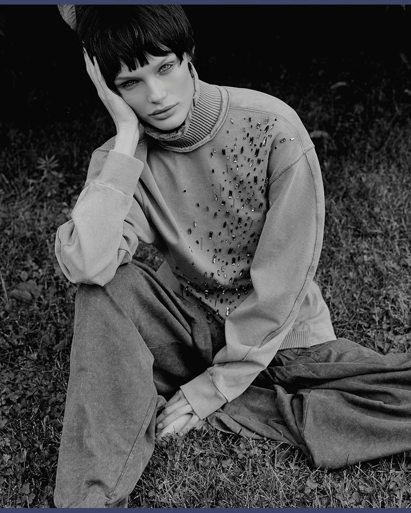 A model with short dark hair sits on grass, wearing a light-colored sweater and dark pants, in a black and white fashion photograph.