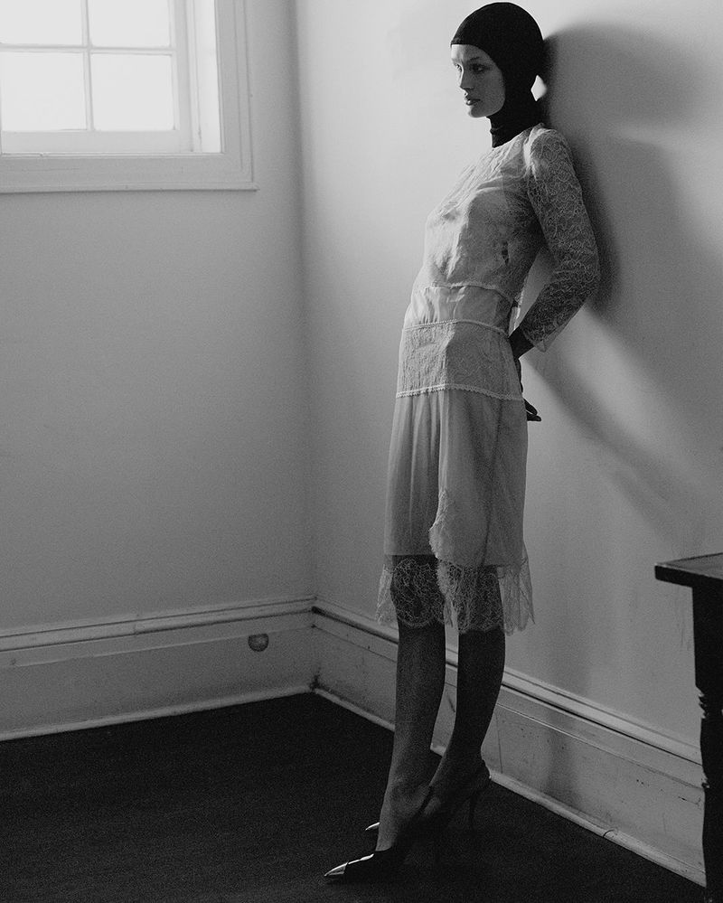 Monochrome portrait of a woman in a lace dress and cap leaning against a wall with a window, offering a stylish and elegant aesthetic.
