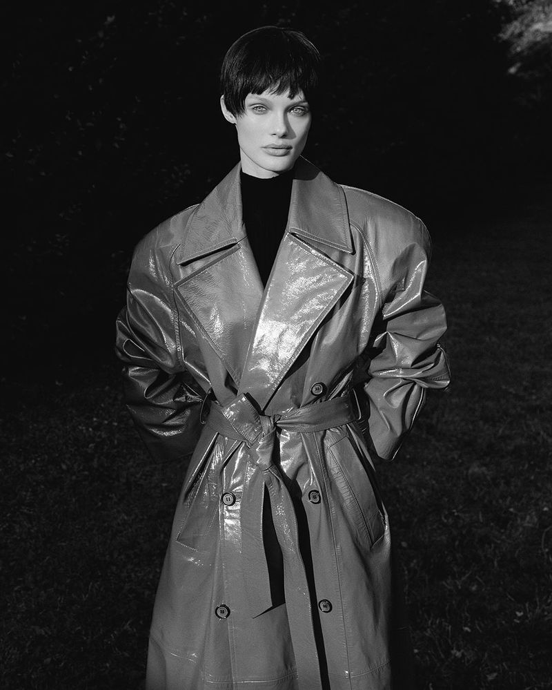 A woman with short dark hair wearing a shiny red trench coat stands in front of a dark background in an outdoor setting.