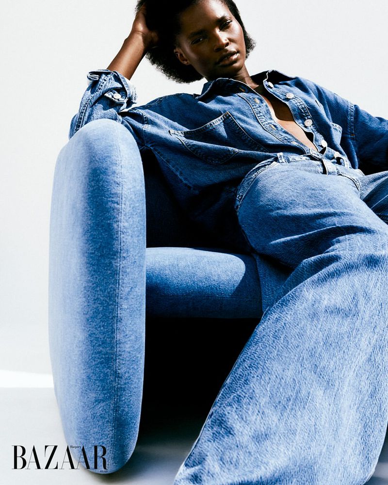 Fashion portrait of a Black female model in a denim shirt and jeans, sitting in a blue chair against a plain white backdrop.
