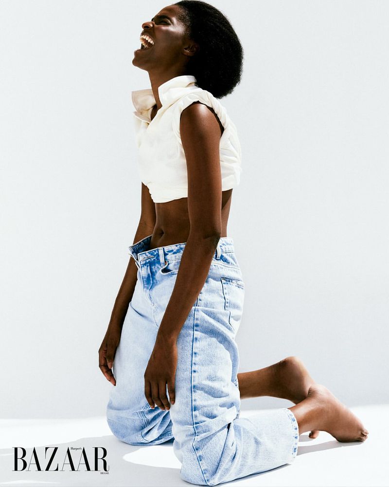 A Black woman kneeling in a white studio, wearing a white sleeveless shirt and jeans, smiling with her head tilted back in laughter.