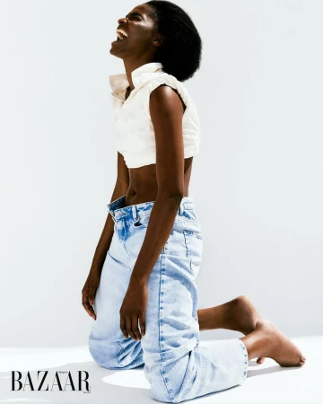 A Black woman kneeling in a white studio, wearing a white sleeveless shirt and jeans, smiling with her head tilted back in laughter.