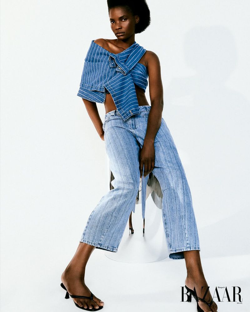 Fashion shot of a Black female model in blue denim two-piece outfit, posing in a photo studio against a white backdrop with black heels.