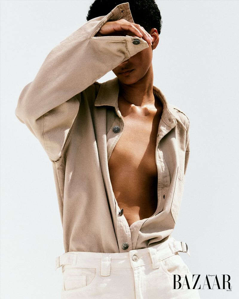 Adult fashion model in a tan button-down shirt, white jeans, and hands covering eyes, posing in a high-key photo studio fashion shot.