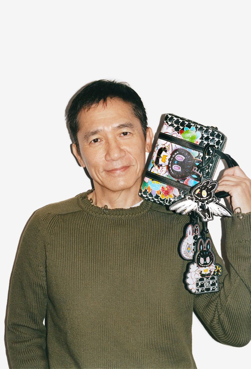 Mature Asian man poses in front of a white background, smiling, holding a colorful patterned bag with cartoon bunny embellishments.