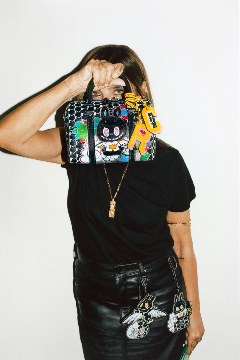 Woman holds a handbag with cartoon-style decorations, wearing a black top and skirt in front of a white backdrop. Fashion shoot.