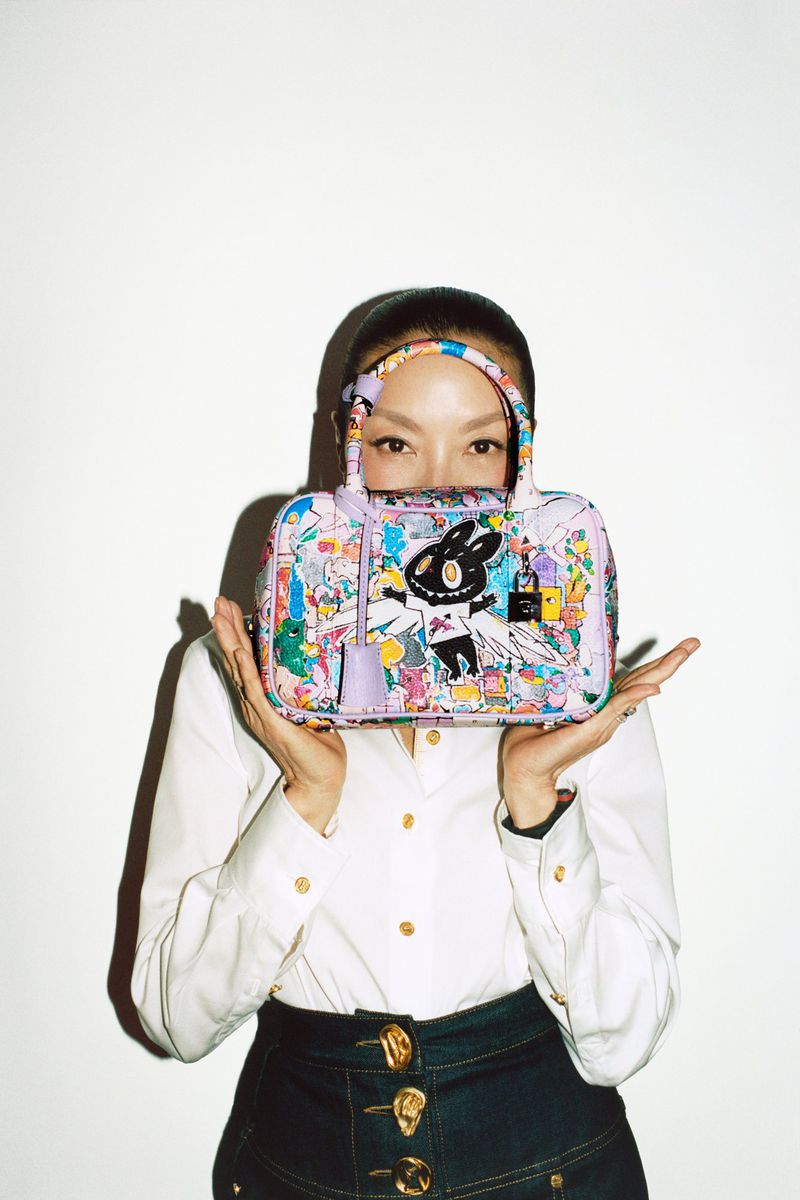 An Asian woman in a button-down shirt and skirt poses with a patterned handbag with cartoon character designs in front of a white wall.