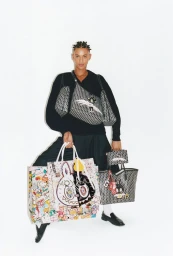 Model wearing a black sweater holding multiple Moynat printed tote bags and wearing crossbody monogram bags against a white background