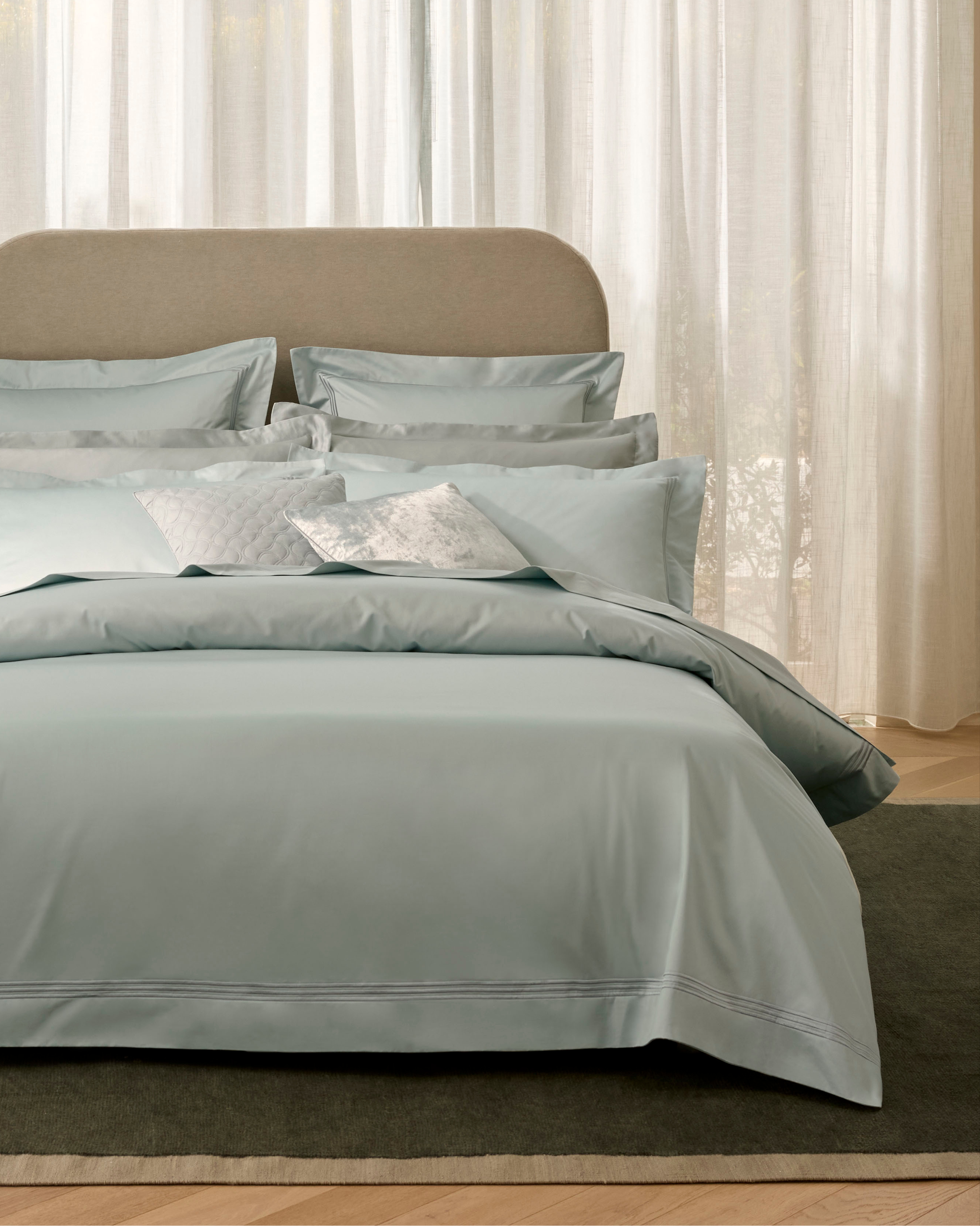 Luxurious bed with light blue bedding, including a duvet cover, pillows, and shams. A neutral headboard complements the cool-toned linen.