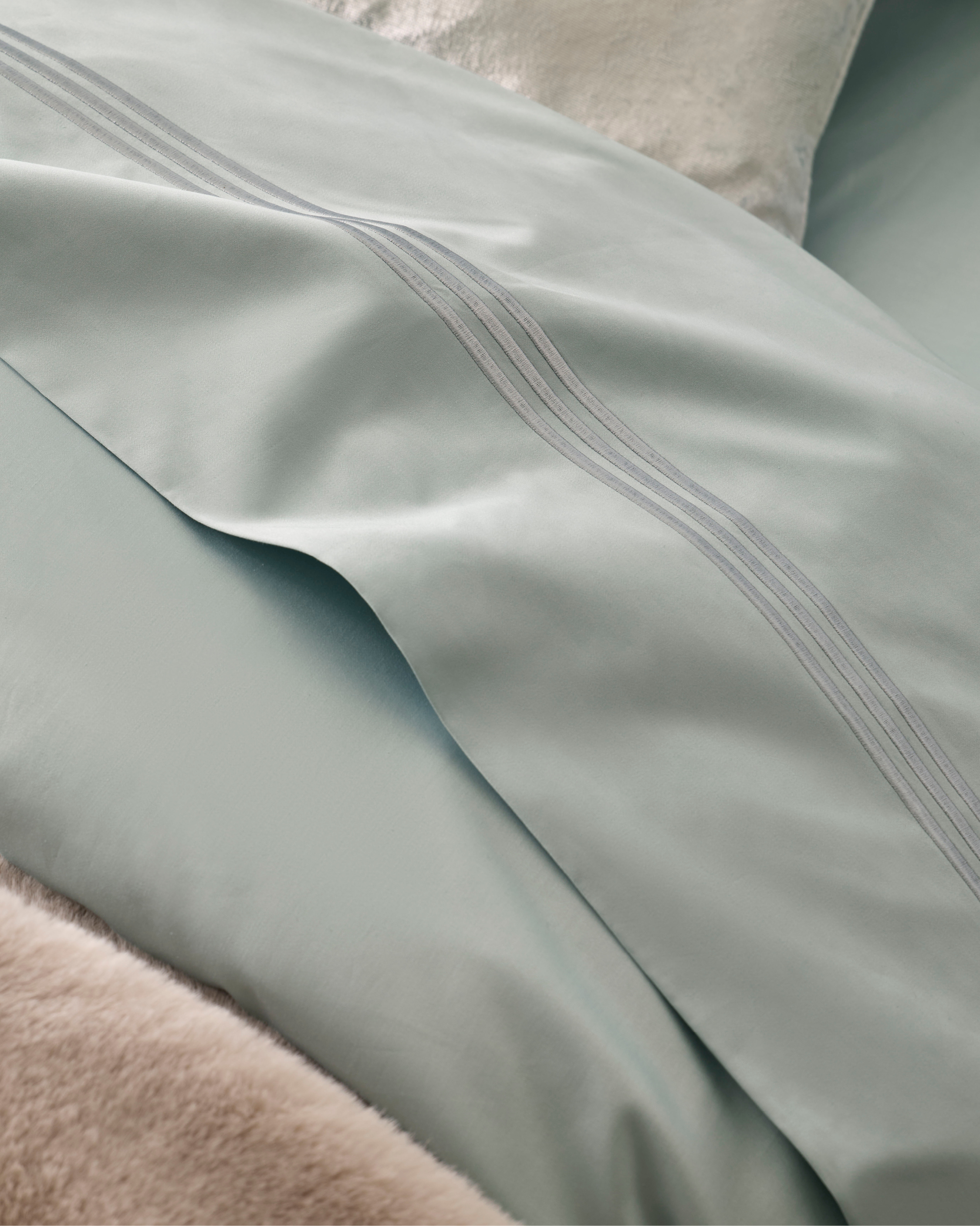 Close-up of light-blue bedding featuring decorative triple-stripe pattern and a soft, neutral-toned textured throw pillow in the background.