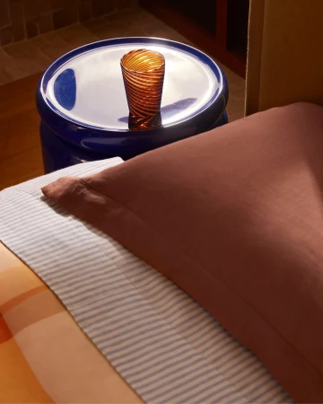 Interior scene features an orange swirled glass on a blue stool next to a bed with striped and orange bedding, creating a cozy and inviting space.