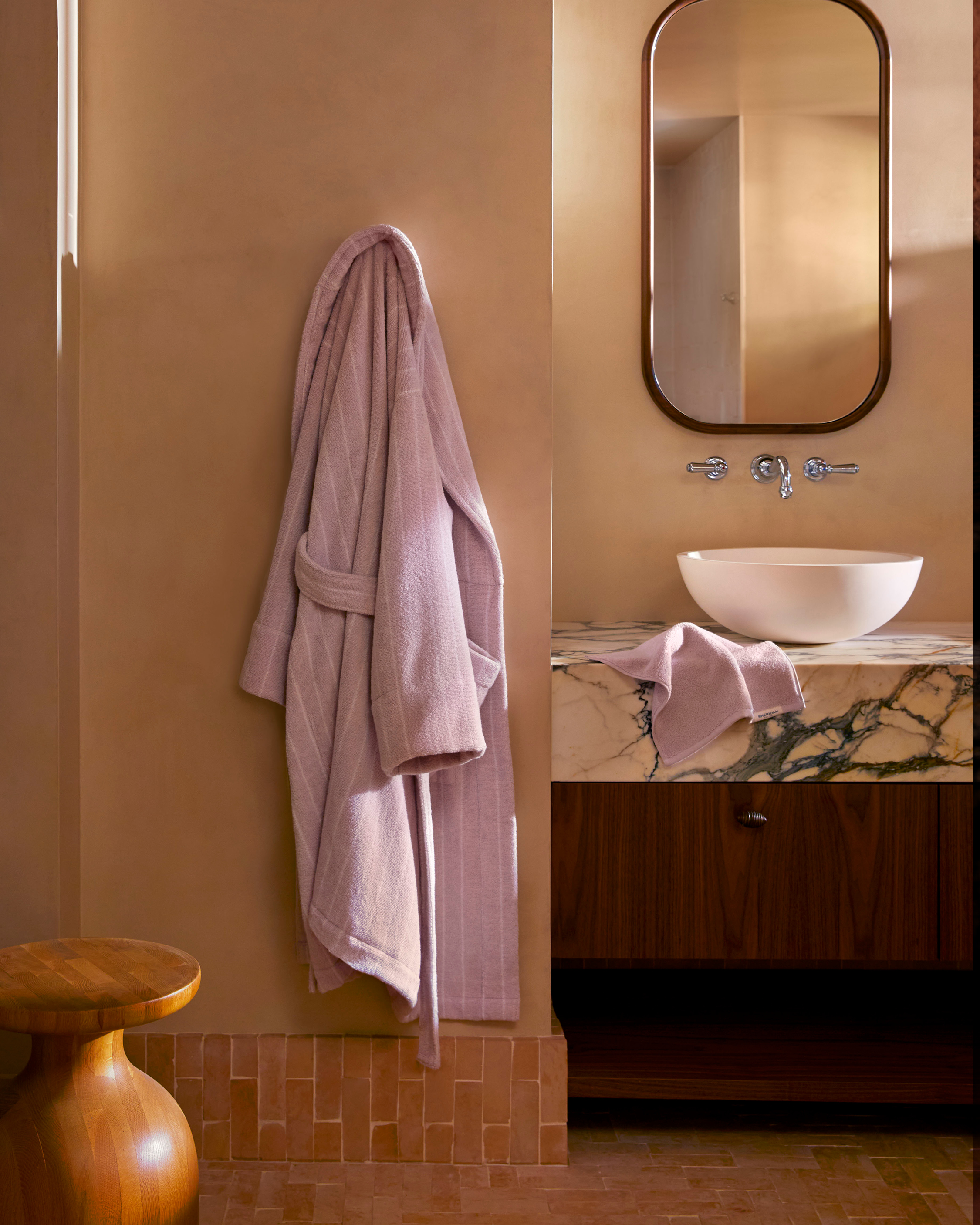 Interior of a bathroom with a light purple striped bathrobe hanging on a tan wall, next to a round mirror and a marble topped vanity.