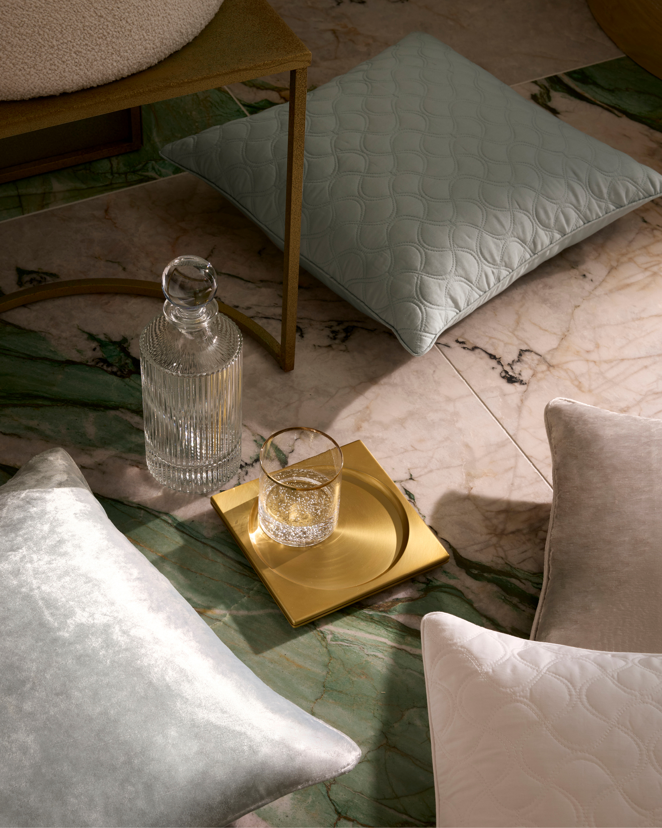 Still life features mint and white decorative pillows, glass decanter, clear glass with liquid, and gold tray on marble floor.