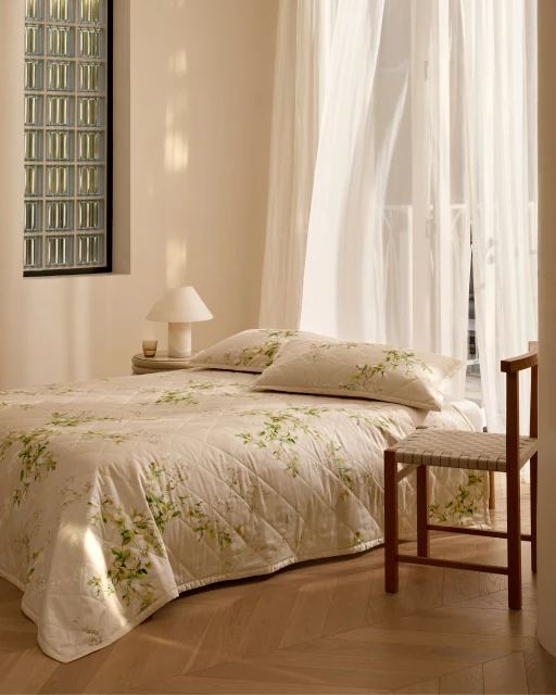 Bright, neutral bedroom interior with a bed featuring floral patterned bedding, a woven chair, and sheer curtains filtering light.
