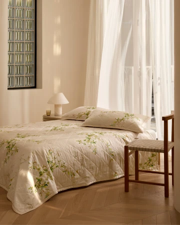 A made double bed with cream patterned bedding and pillows beside a wooden chair in a sunlit bedroom.