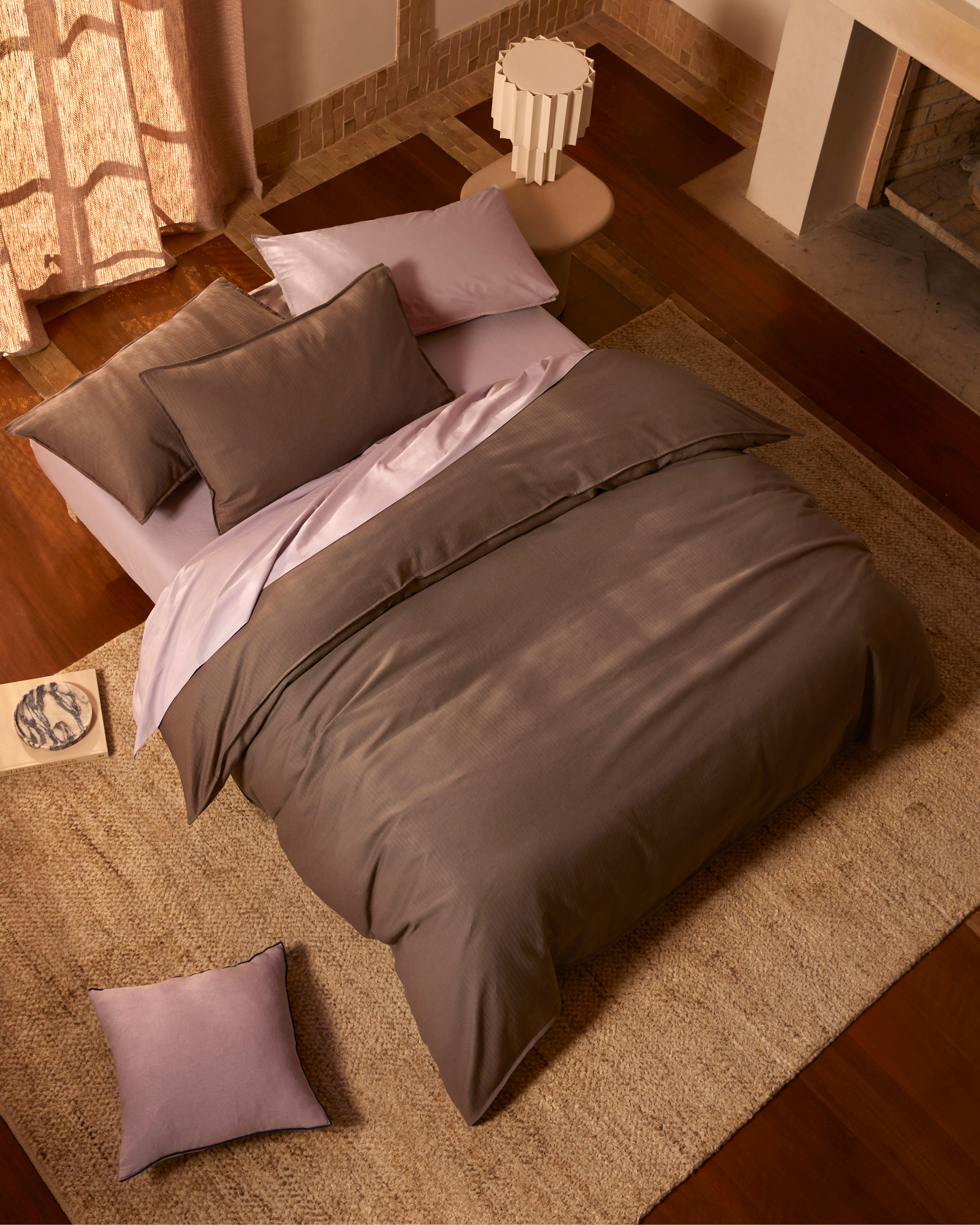 High-angle shot of a neatly made bed with brown duvet and lavender sheets, complemented by a soft beige rug in the bedroom.