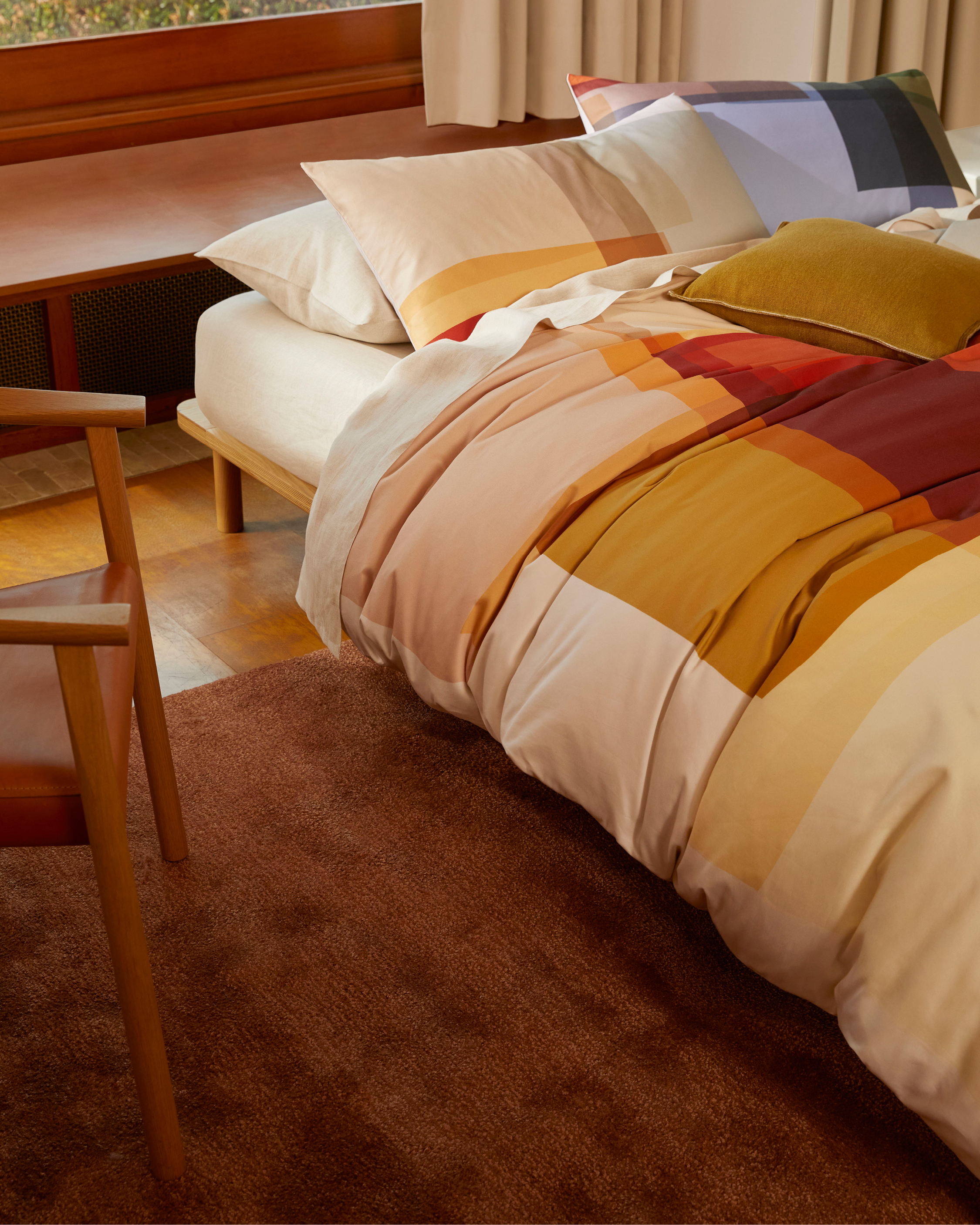 Bedroom interior featuring a bed with patterned bedding, pillows, wooden headboard, natural light from a window, and a wooden chair.