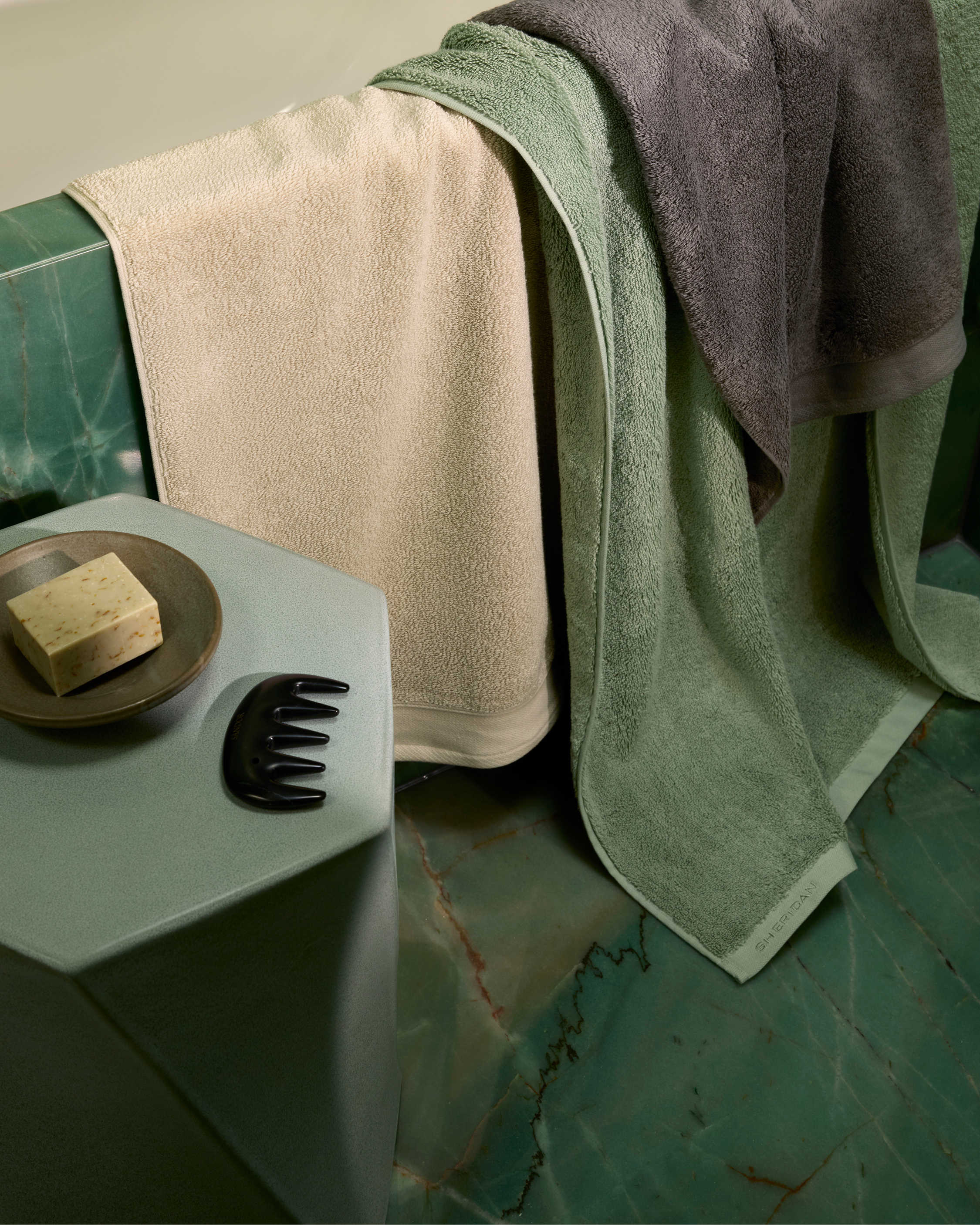 High-angle view of neatly folded beige, green, and gray towels hanging over a green marble bathtub, with a comb and soap.