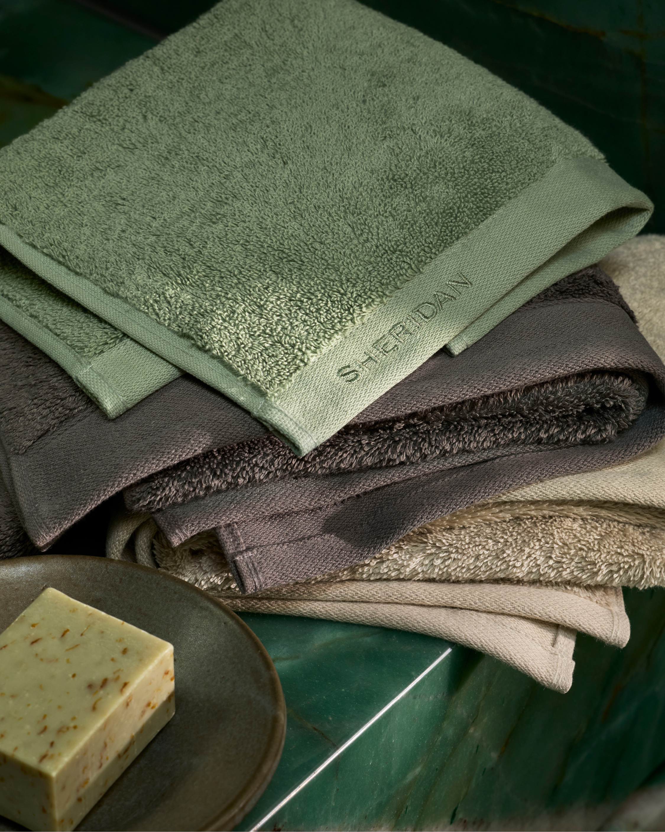 Close-up of folded towels in shades of green, gray, and beige, next to a bar of soap on a green marble surface.