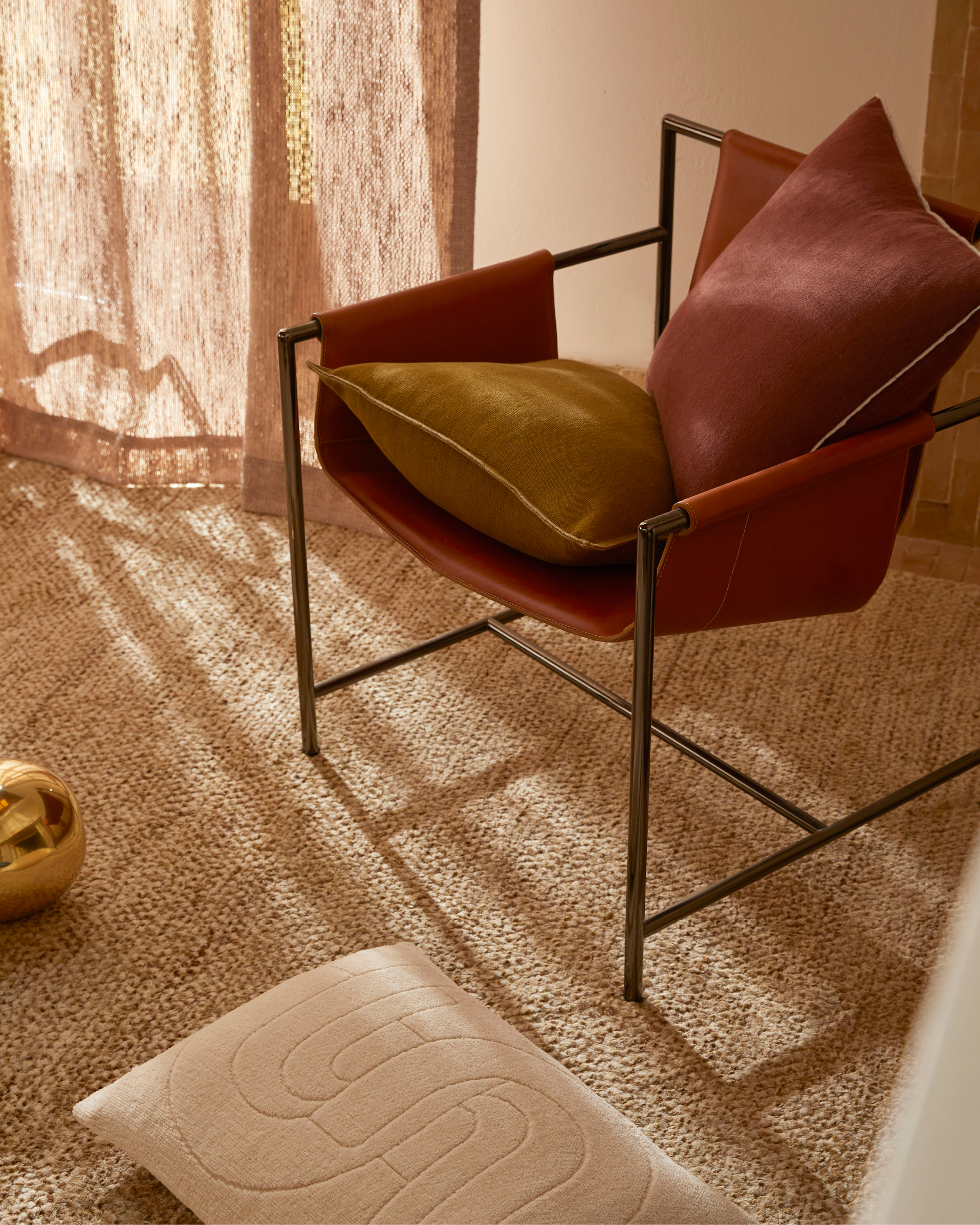 Modern interior: armchair with cushions on a textured rug near a window with neutral-toned curtains, creating a cozy aesthetic.