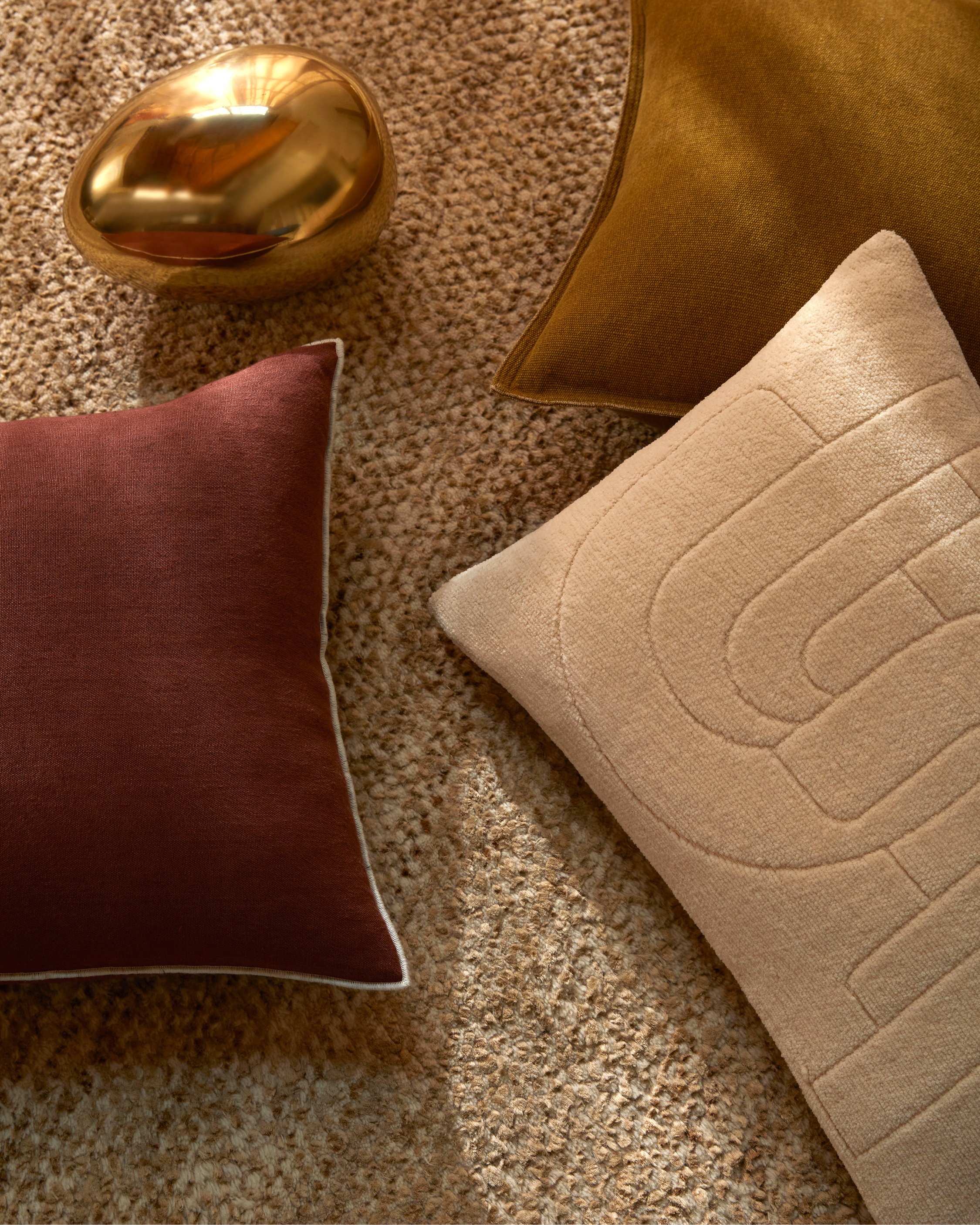 Still life featuring throw pillows in shades of brown, tan, and maroon, arranged on a beige textured carpet, with a golden decorative sphere.