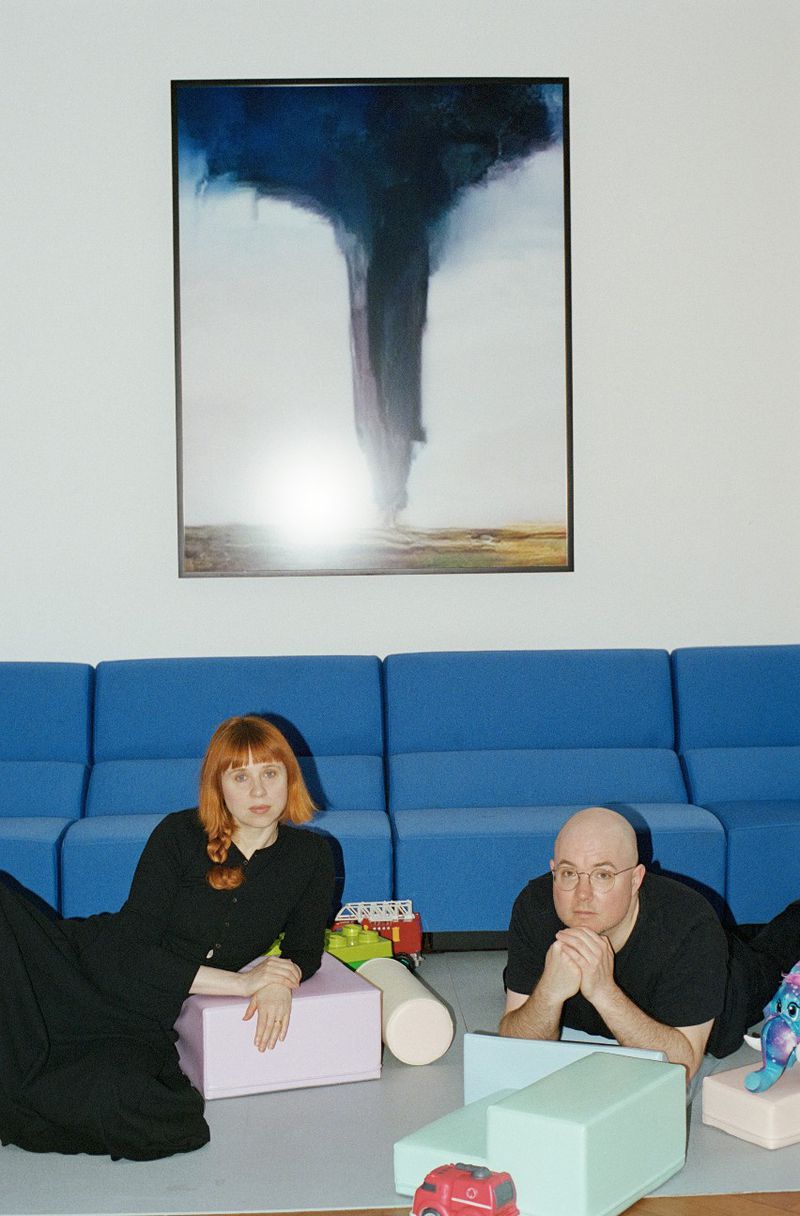 Woman with red hair and a man lying on the floor surrounded by colorful foam blocks and toys, in front of a blue couch and art.