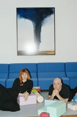 Woman with red hair and a man lying on the floor surrounded by colorful foam blocks and toys, in front of a blue couch and art.
