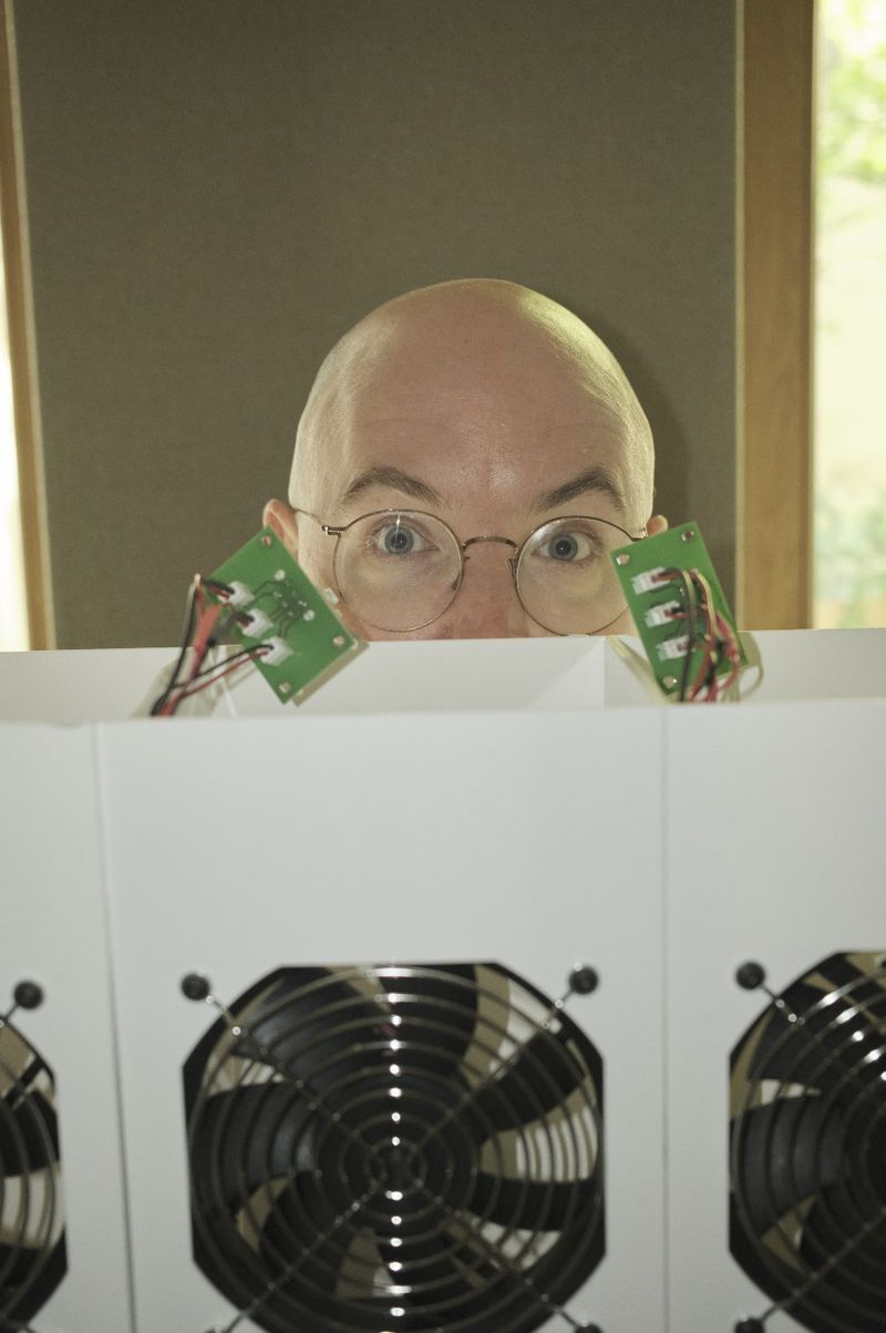 A bald adult peers over a white barrier alongside green circuit boards, positioned in front of cooling fans in an indoor setting.