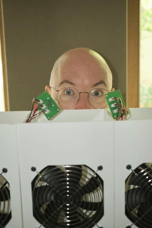 Bald person wearing round wire-rimmed glasses peeking over white computer hardware with visible cooling fans.