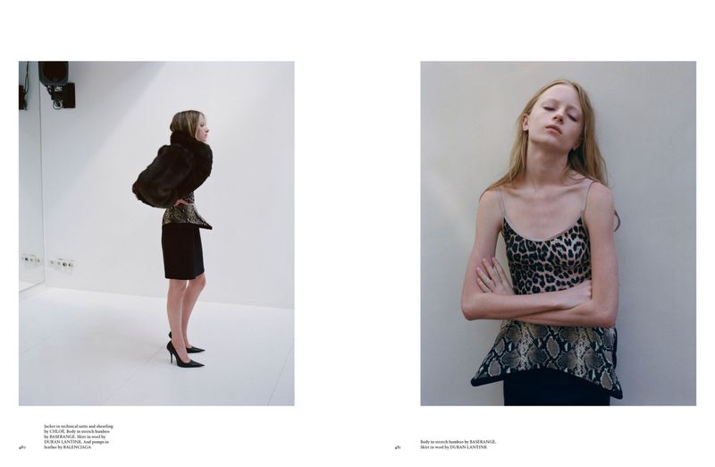 Fashion editorial featuring a blonde woman wearing a black dress and fur coat, indoors with a white wall background. Another woman wears a leopard print top.