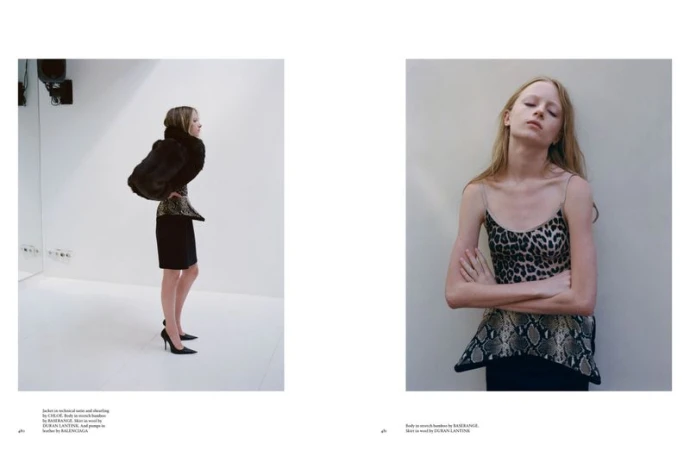 Fashion editorial featuring a blonde woman wearing a black dress and fur coat, indoors with a white wall background. Another woman wears a leopard print top.