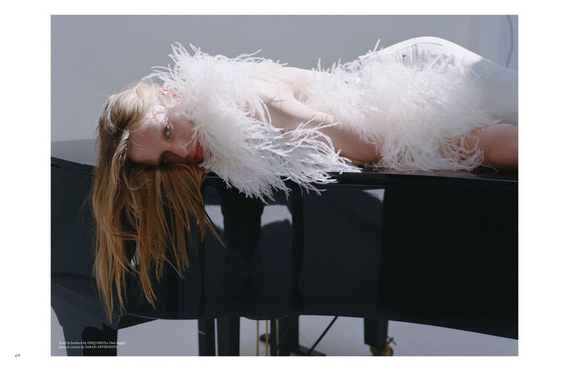Woman in feathered dress lies on grand piano indoors. Beauty shot with fair skin, long blonde hair, and dark glossy piano.