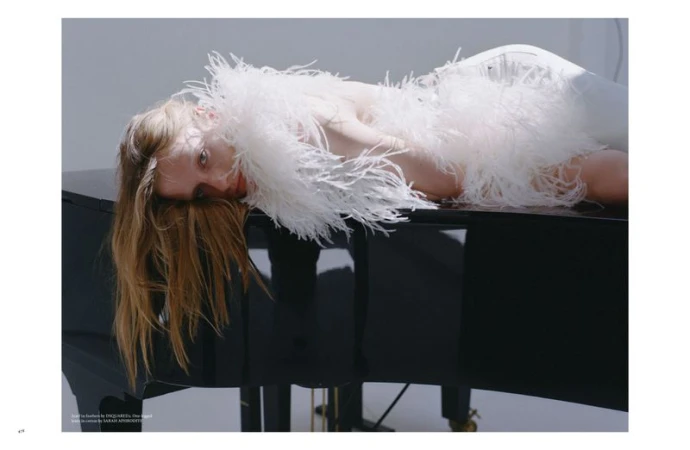 Woman in feathered dress lies on grand piano indoors. Beauty shot with fair skin, long blonde hair, and dark glossy piano.