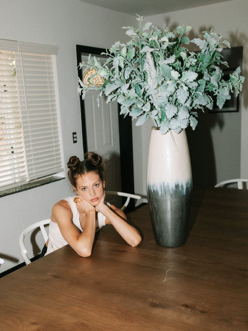 Young woman with hair in space buns sits at a wooden table with a vase of leafy foliage in a domestic interior setting.