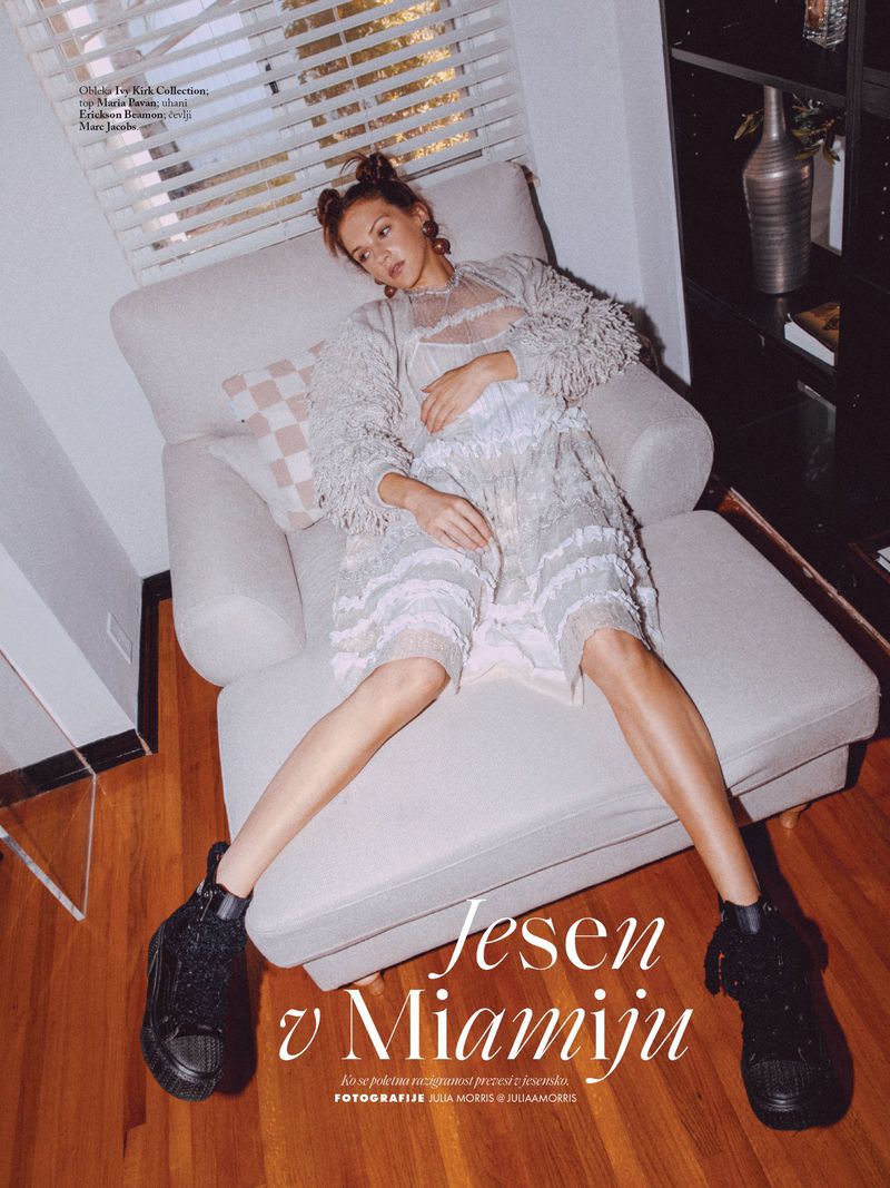 Fashion editorial shot of a woman in a dress and sweater on a chaise lounge with black boots, near a bookcase and a window with blinds.